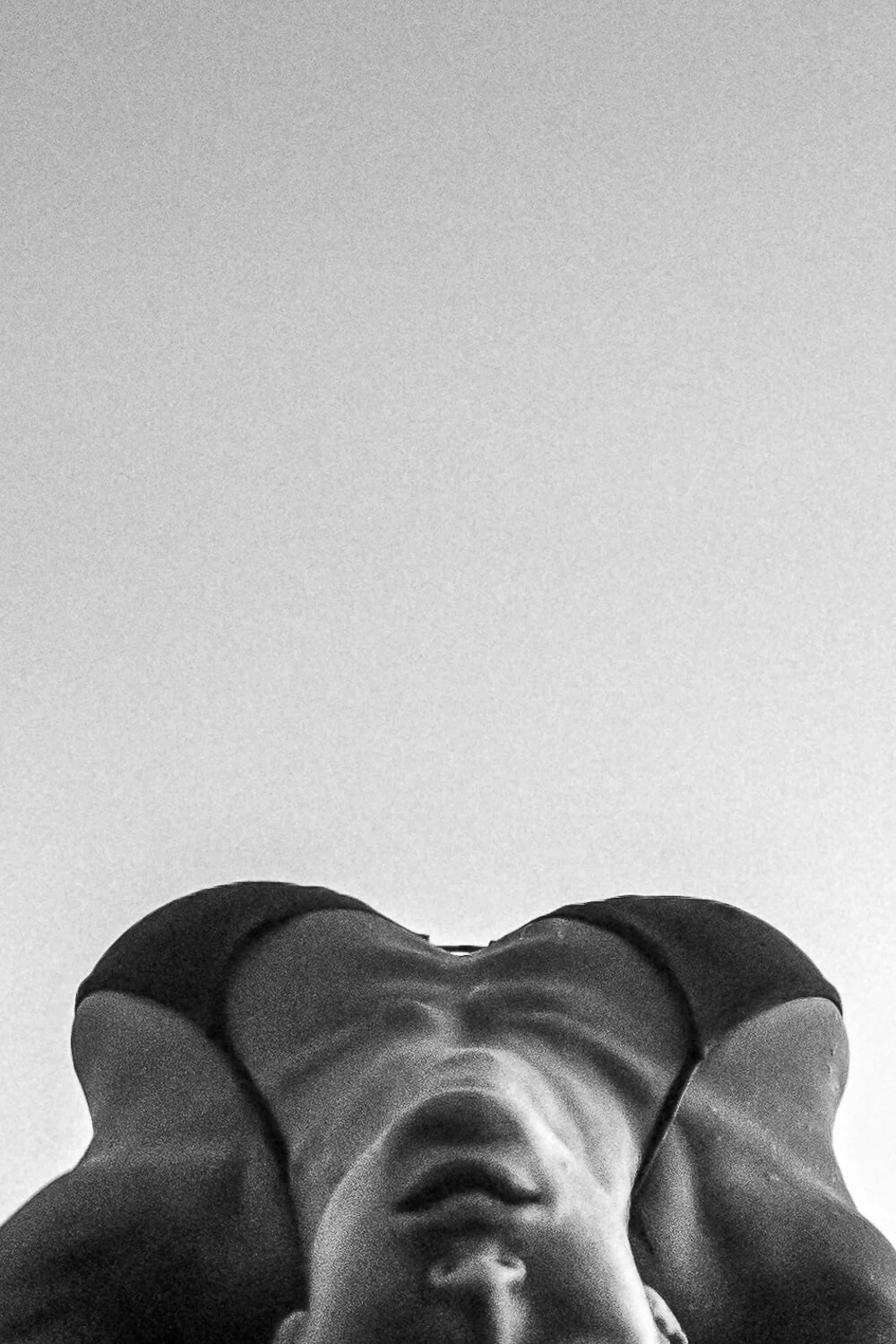 Black and white photo of a person lying down on their back with their hands behind their head, viewed from below, with a plain white ceiling in the background.