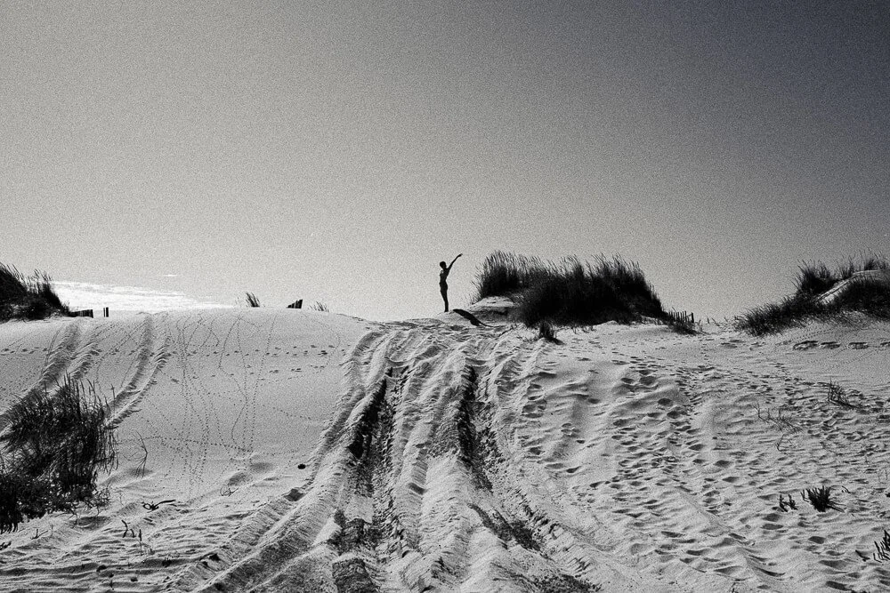 A person standing on top of sand dunes with tire tracks and footprints, surrounded by grassy vegetation, under a clear sky.