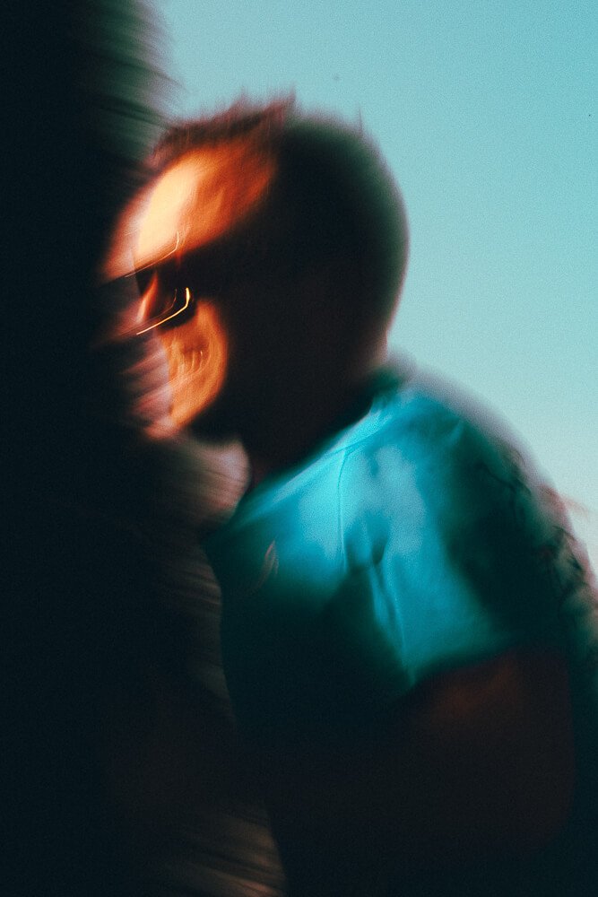 Blurred photo of a person with short hair and glasses, wearing a light blue shirt, against a plain background.