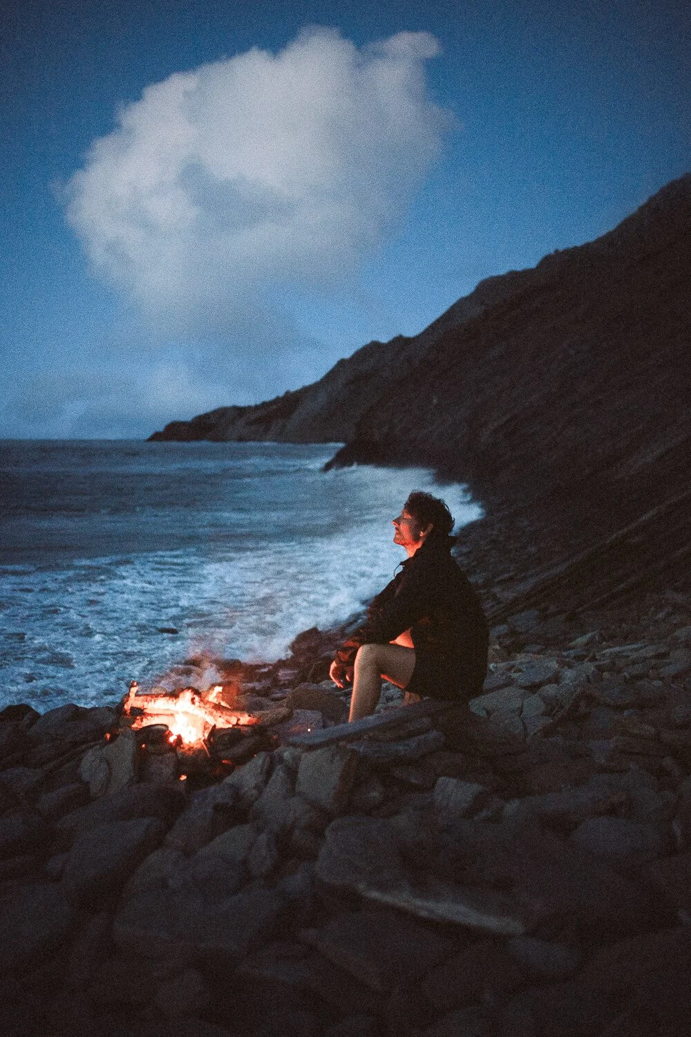 Person sitting on a rocky beach at dusk, near a small campfire, with rugged cliffs and ocean waves in the background.