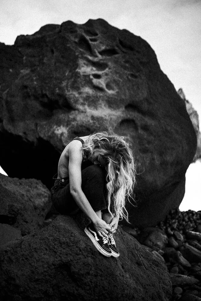 A woman with long curly hair sitting on rocks near a large boulder, with her head bowed and hands adjusting her shoes, in a grayscale outdoor setting.