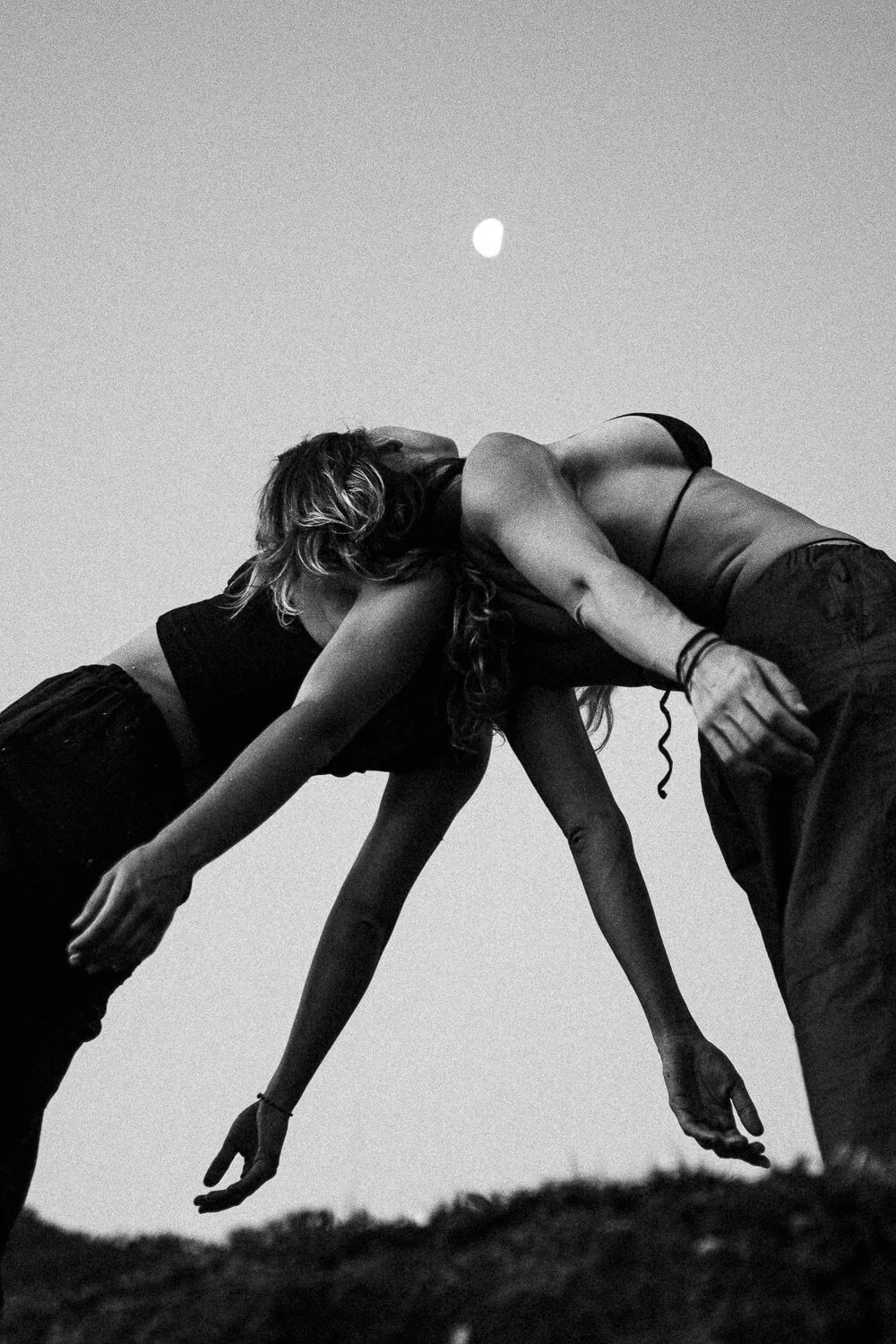 Two women are performing a yoga pose outdoors during sunset or dusk, with the moon visible in the sky above.