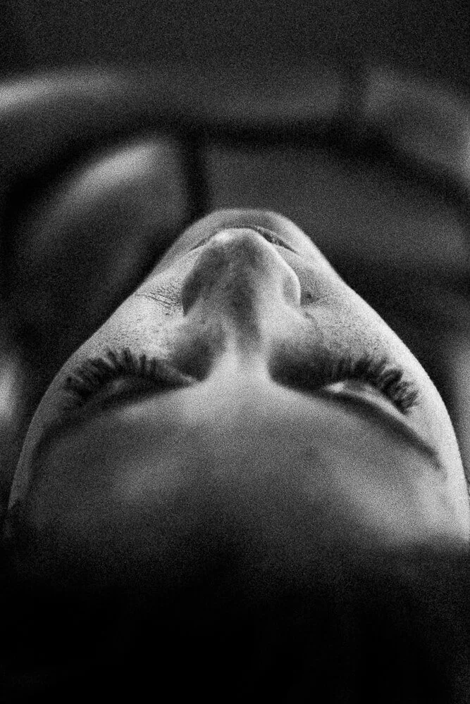 Close-up of a person's face as they look upwards, with long eyelashes and detailed facial features, in black and white.