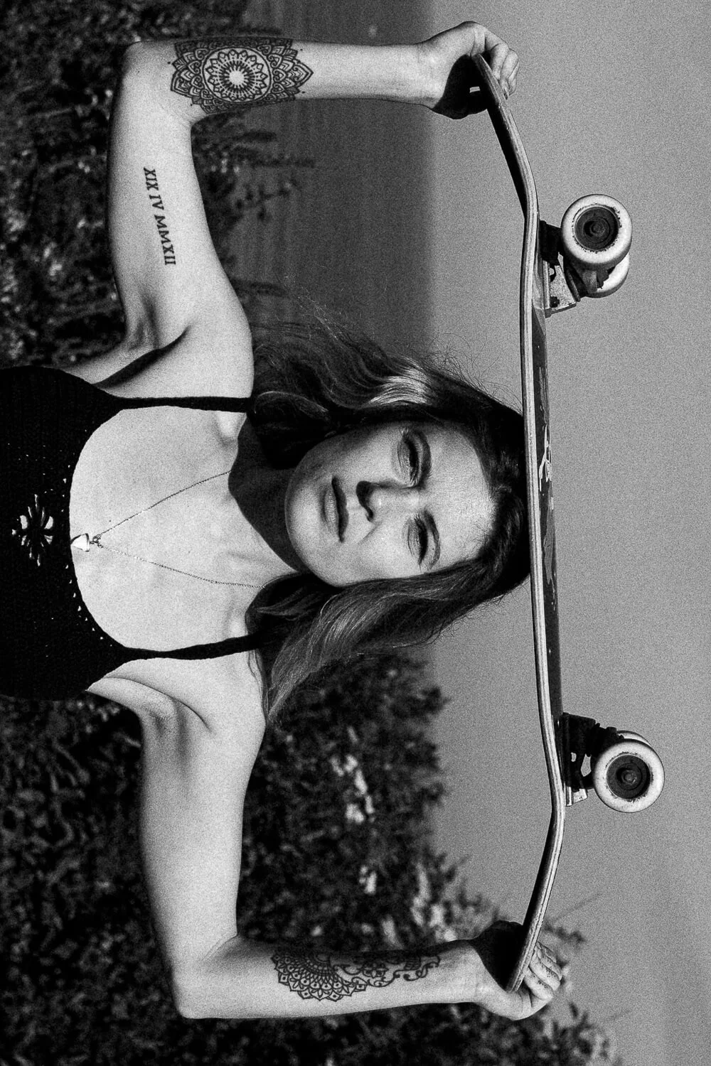 A woman with tattoos on her arms and a necklace, lying on the ground outside with a skateboard above her head.