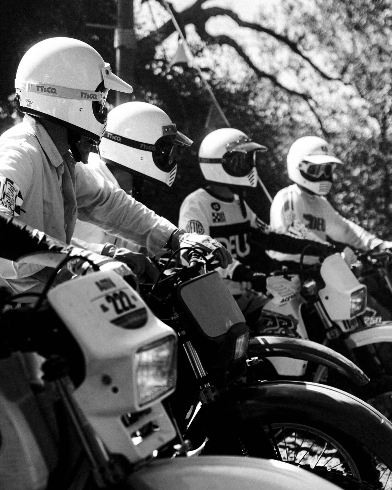 Line of motorbikes with helmets resting on the handlebars, outdoors with trees in the background.
