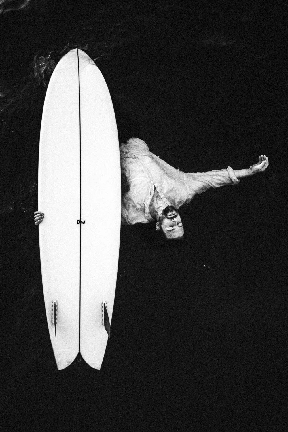 A man in a light-colored shirt and sunglasses is lying on his back in dark water, holding a surfboard vertically next to him, with his head and part of his torso visible.