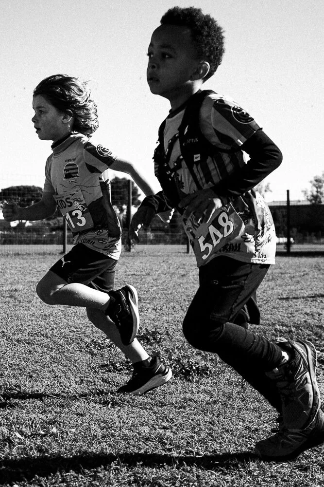 Two children running on a dirt field during a race, wearing athletic clothing and numbered bibs.