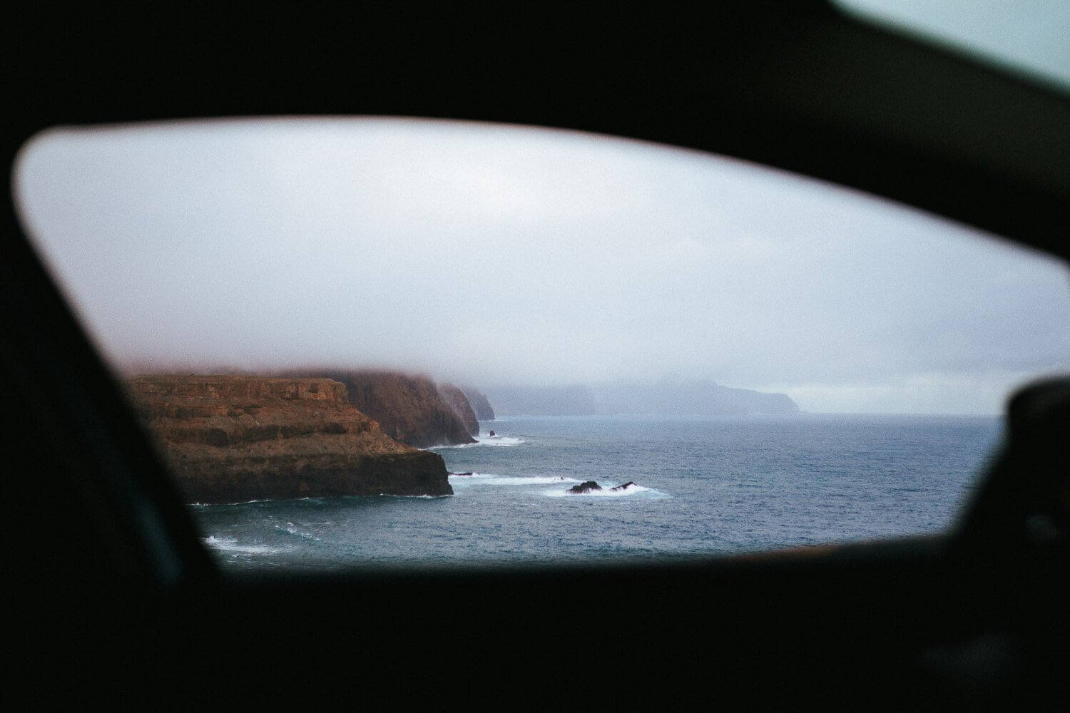 Cliffs along a rugged coastline view through a car window with ocean waves and cloudy sky