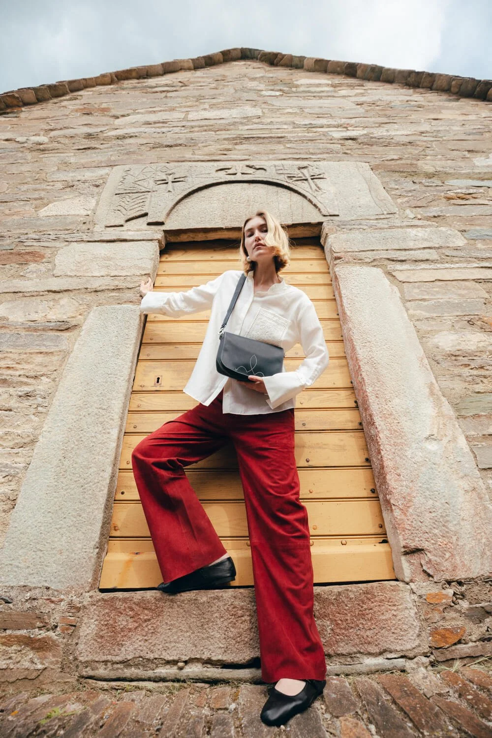 A young woman with blonde hair wearing a white blouse and red pants, standing in front of a stone wall with an arched wooden door, looking down at the camera.