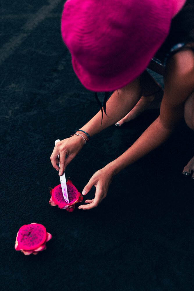 Person with a pink hat slicing a pink dragon fruit with a knife on a black surface.