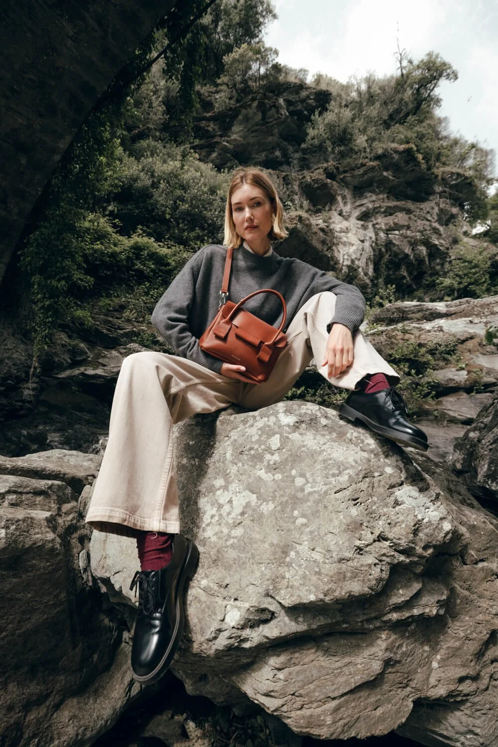 A woman sitting on a large rock outdoors with a forested hillside behind her, dressed in a gray sweater, beige pants, black shoes with burgundy socks, holding a brown handbag.
