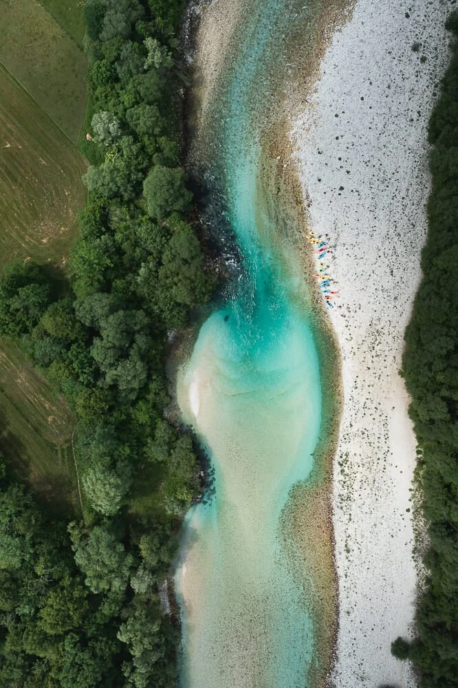Aerial view of a turquoise lake surrounded by green trees and a rocky shoreline, with people on the beach near the water.