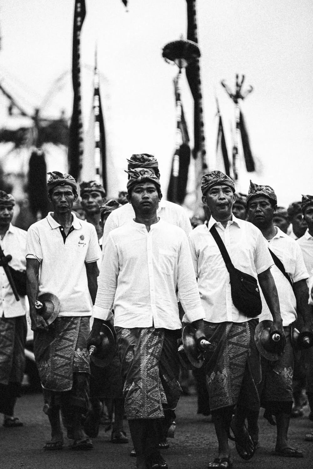 Groupe de personnes en marche portant des vêtements traditionnels et des instruments de musique, probablement lors d'un festival ou d'une parade culturelle, avec des drapeaux en arrière-plan.
