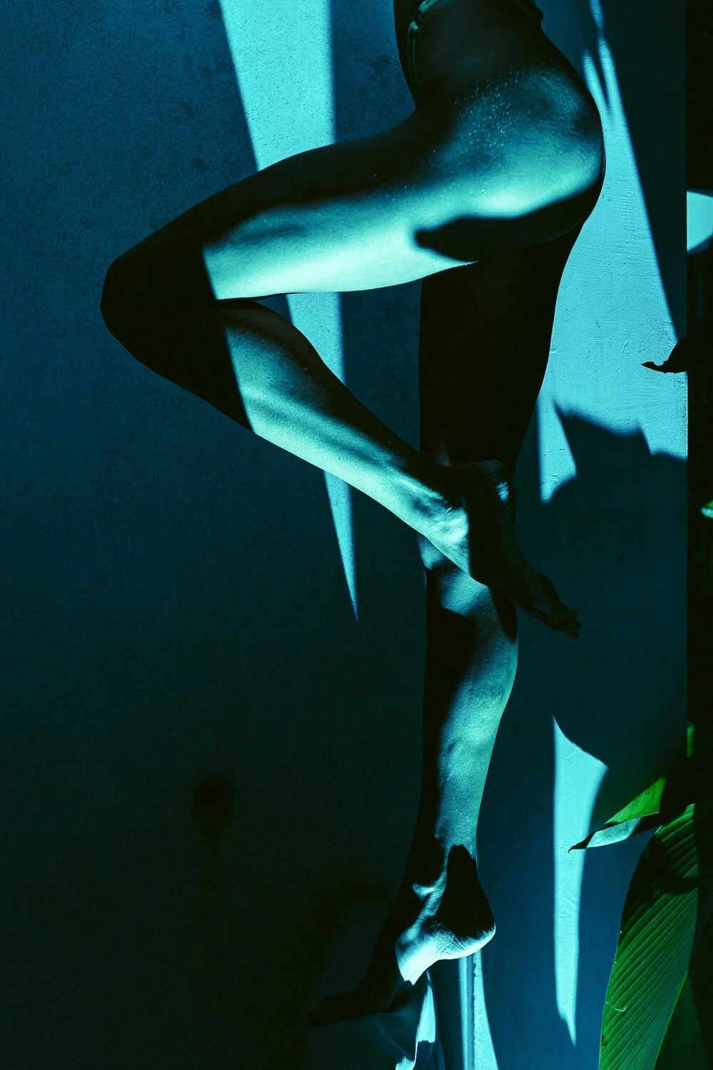 An abstract photograph of a person's arm and leg, cast in blue-green lighting, with strong shadows and a textured wall background.