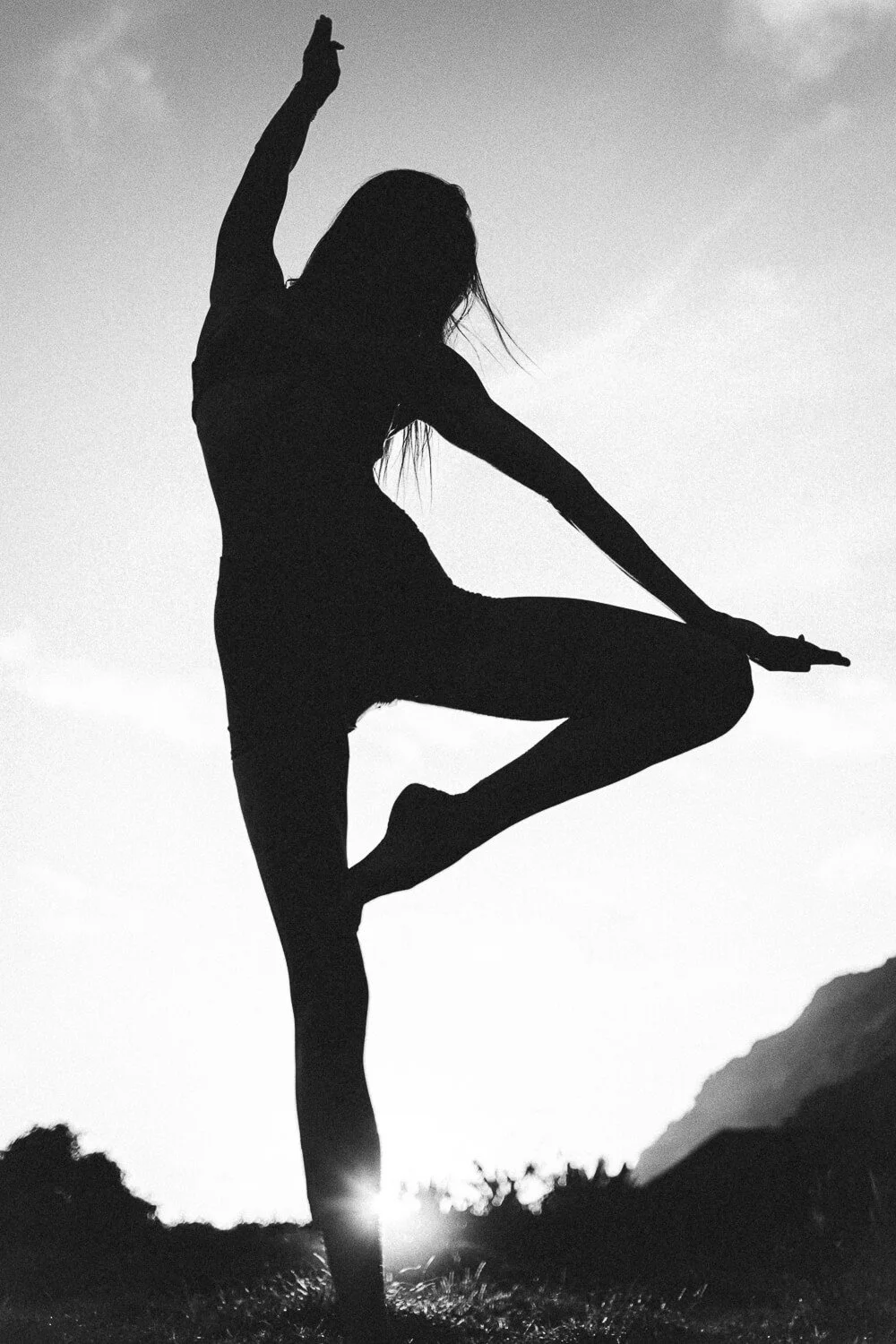 Silhouette of a person practicing yoga outdoors during sunset or sunrise, with mountains in the background.