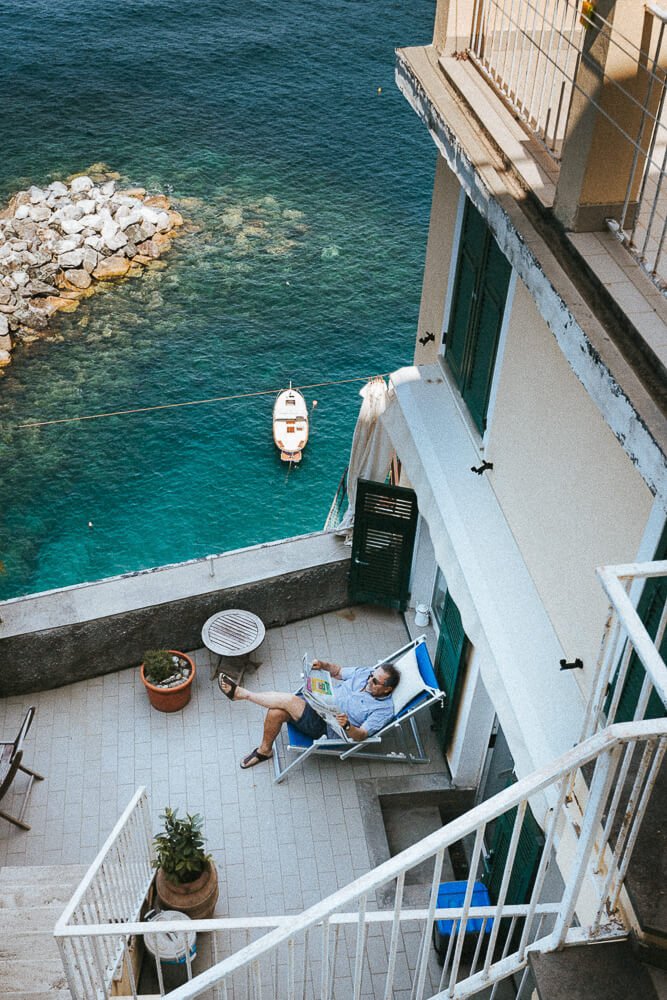A man relaxing on a balcony over the water, reading a newspaper, with a small boat floating nearby.
