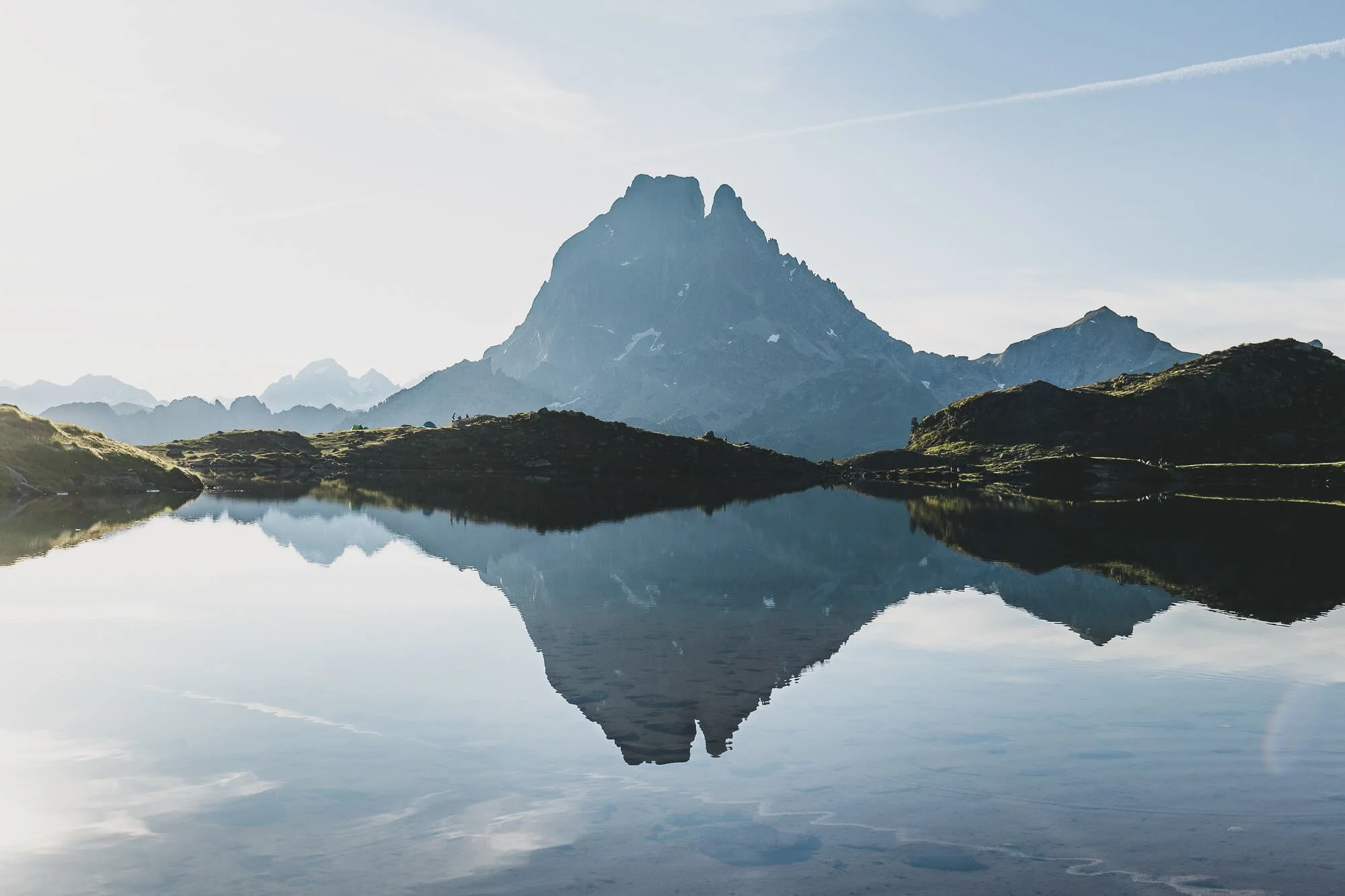 A mountain reflected in a calm lake, with grassy hills and a light blue sky in the background.