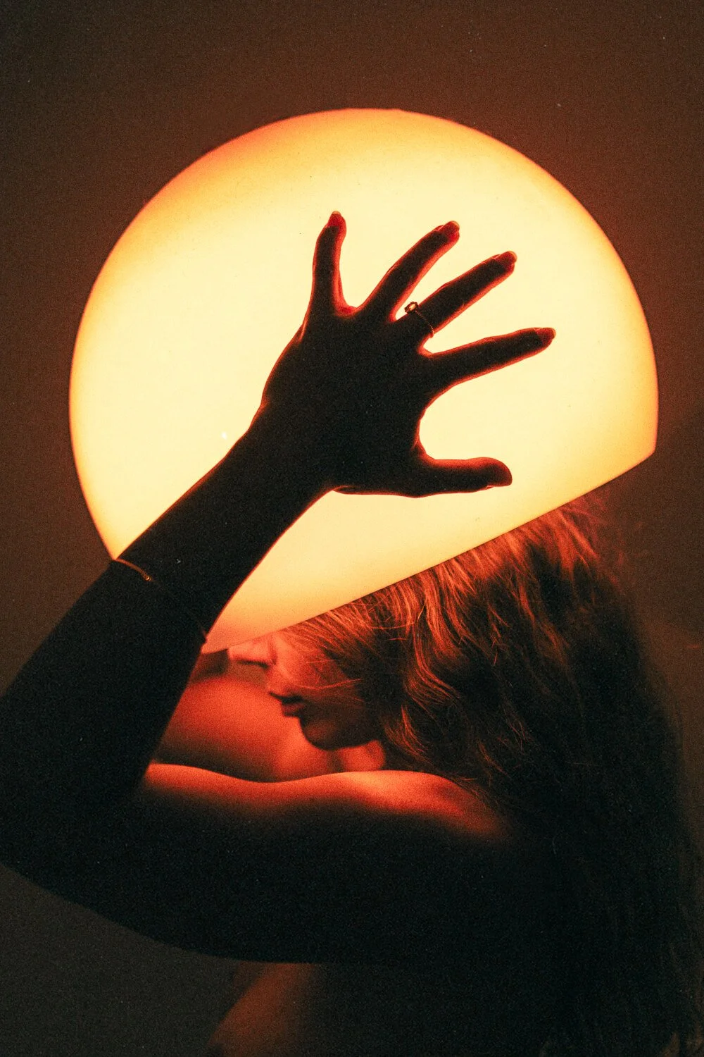 A person with long hair and a dark long-sleeve shirt holding a large glowing lamp over their head, with their face turned down and partially obscured.