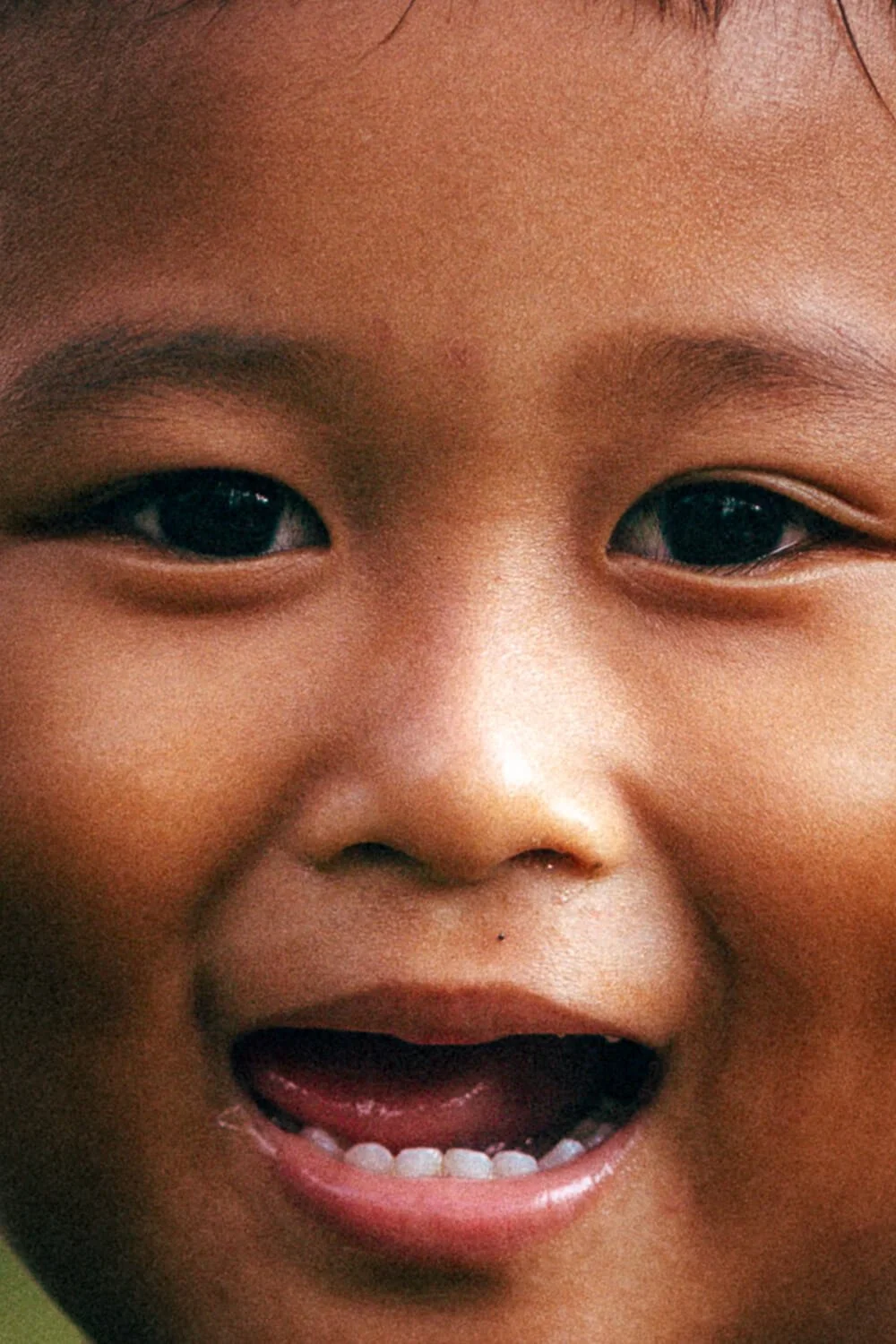 Un gros plan du visage souriant d'un enfant asiatique, montrant ses yeux, son nez, sa bouche ouverte et quelques dents.