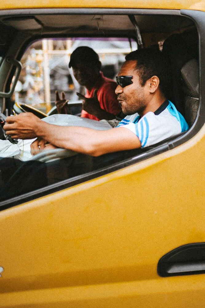 Two men sitting in a yellow triangle-shaped vehicle. The man in front is wearing sunglasses and a blue and white shirt, smiling and looking to his right. The man in the back is wearing a red shirt and gesturing with his hand. The vehicle appears to b