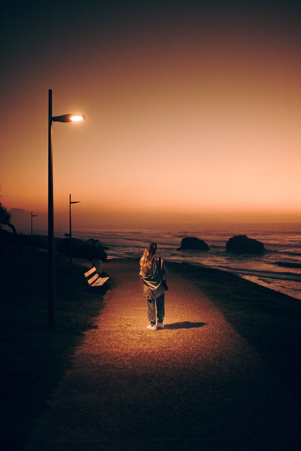 A woman walking along a pathway by the ocean at sunset, illuminated by streetlights with a pink and orange sky.