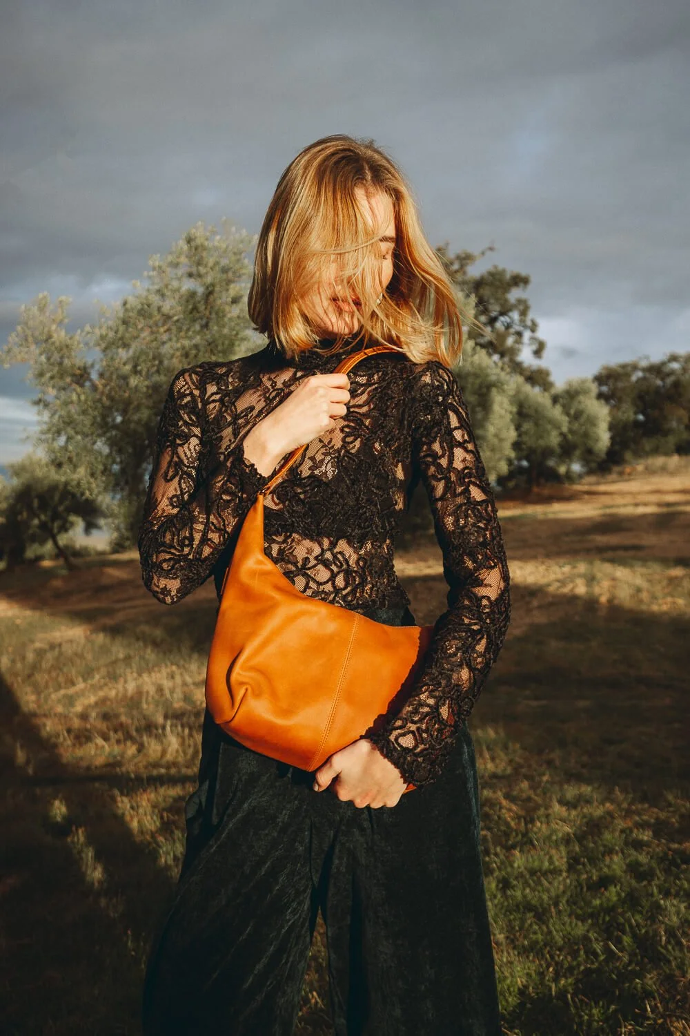 Woman with shoulder-length blonde hair wearing a black lace top and black pants, holding a tan leather shoulder bag, standing outdoors on a grassy field with trees and a cloudy sky in the background.