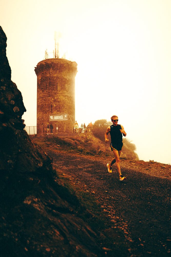 A person running on a dirt trail near an old stone tower with a sign that says 'ARRIVÉE' at the top of a mountain or hill during sunset or sunrise.