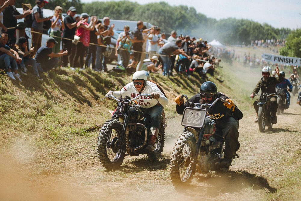 Two motocross racers in vintage bikes, one wearing white and the other black, racing on a dirt track with a crowd of spectators watching from the sidelines.