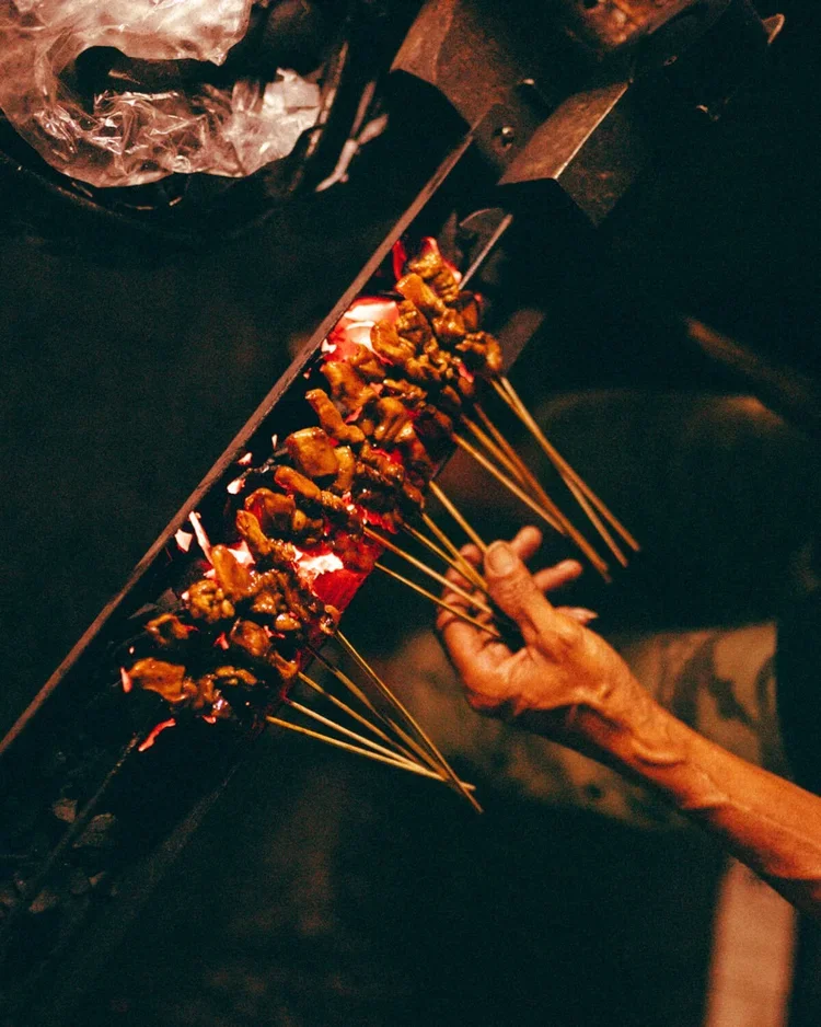 Une main tenant des brochettes de viande grillée sur un barbecue dans un environnement sombre.