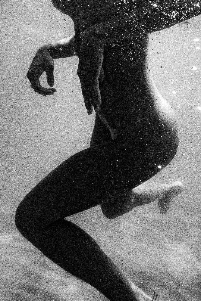 Black and white underwater photo of a nude woman with her hand on her thigh, swimming in the water.