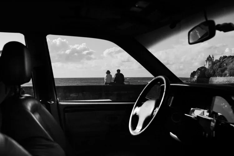 Vue depuis l'intérieur d'une voiture en noir et blanc, avec deux personnes assises au bord de la mer, regardant l'horizon, et un château sur une colline en arrière-plan.