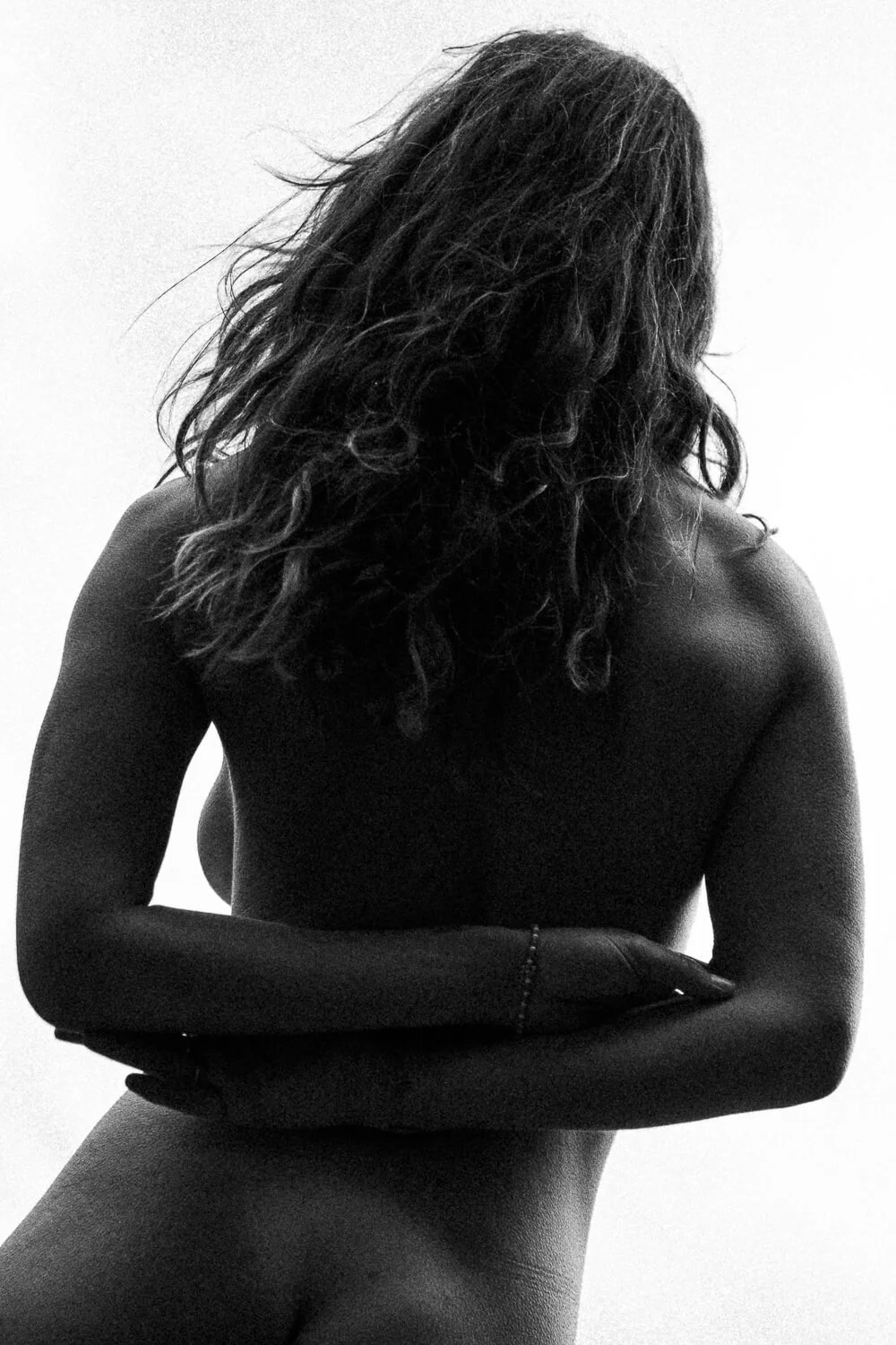 Black and white photo of a nude woman with curly hair, her arms crossed over her chest, and her face turned away from the camera.
