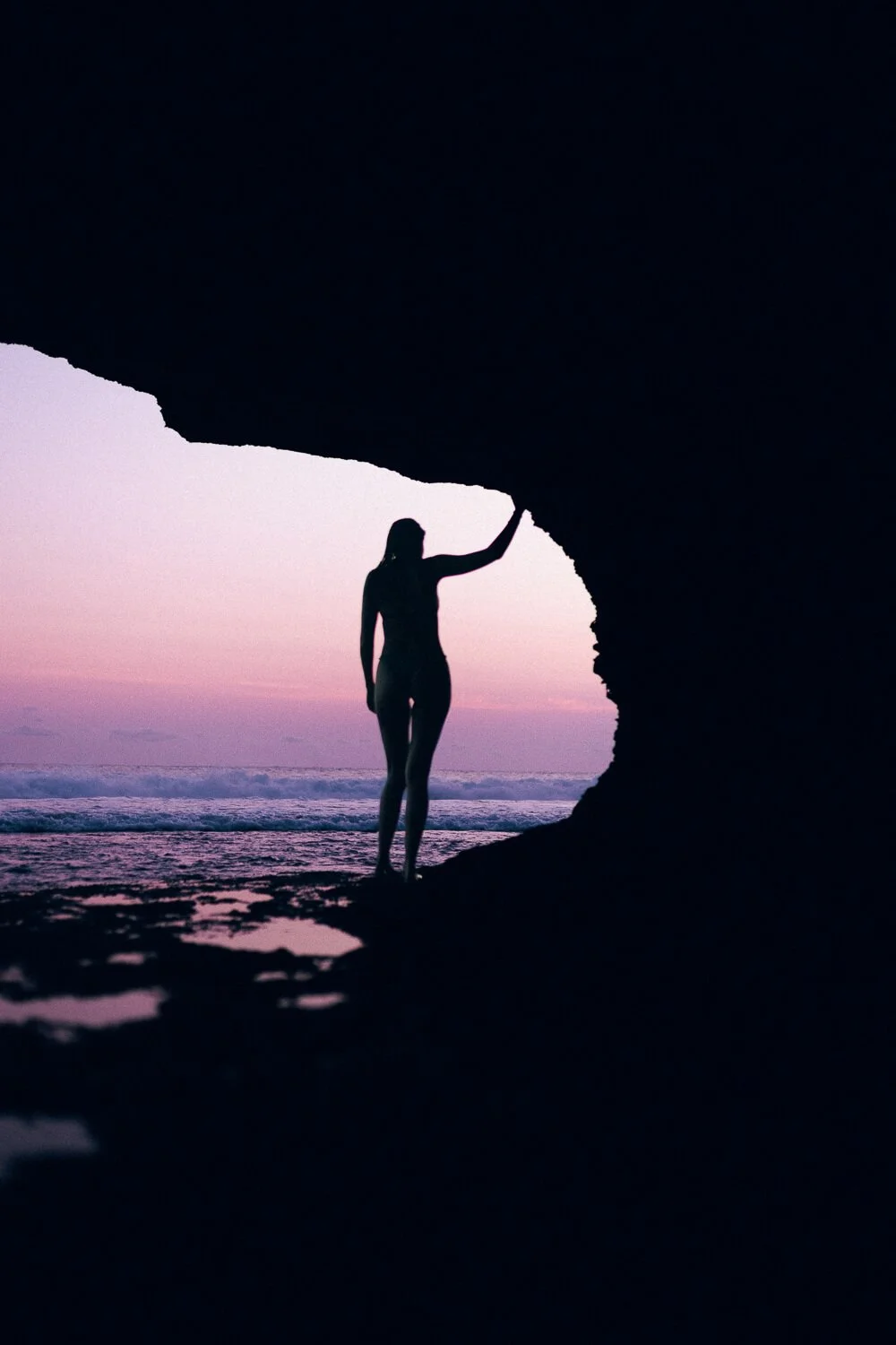 Silhouette of a woman standing under a rock formation at the beach during sunset, with waves in the background and pink and purple sky colors.