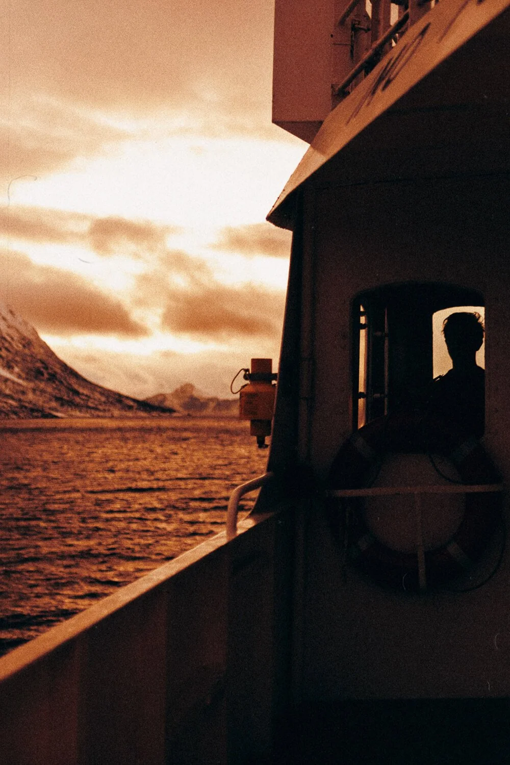Silhouette d'une personne dans la cabine d'un bateau naviguant près de montagnes au crépuscule, ciel orange.
