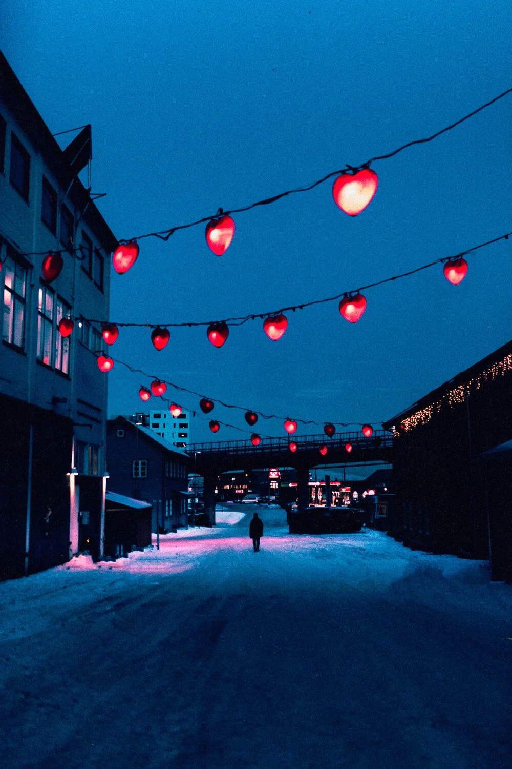 Des guirlandes lumineuses en forme de cœurs rouges suspendues dans une rue enneigée au crépuscule, avec une silhouette d'une personne marchant au centre.