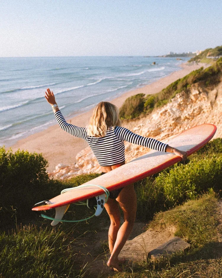 Une femme avec une planche de surf, en robe à rayures, marchant vers la plage à côté d'une falaise verdoyante, avec l'océan en arrière-plan.