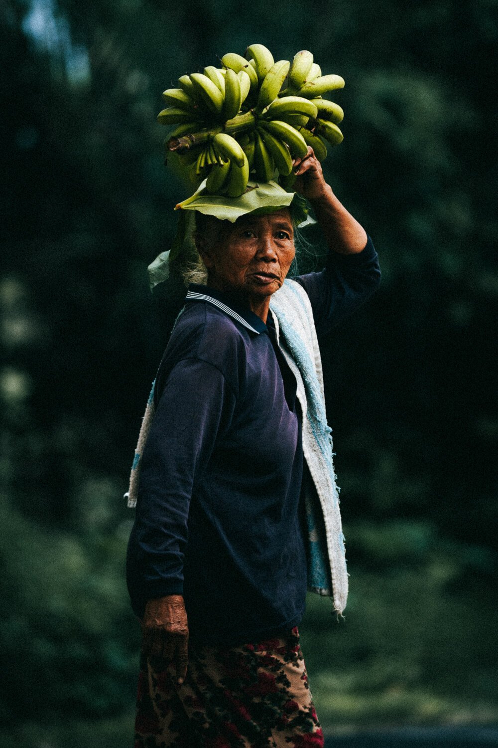 Une femme âgée portant un panier de bananes sur sa tête, dans un environnement naturel.