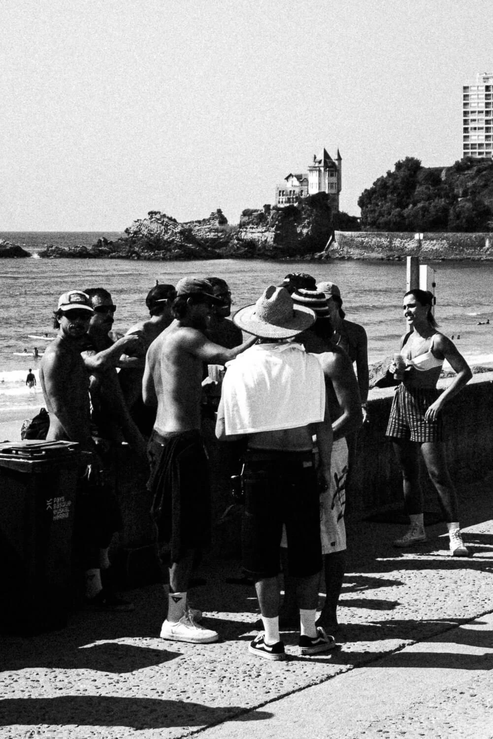 Group of people gathered on a beach in front of a shoreline with houses and trees in the background, sunny day, black and white photo.