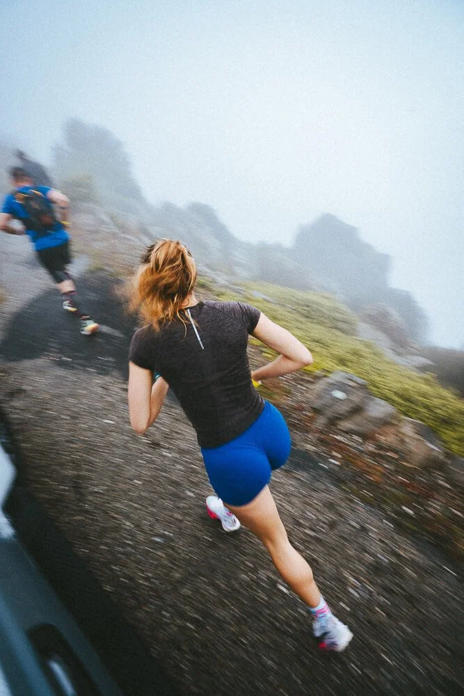 Two women running uphill on a foggy, rocky trail in a mountainous area.
