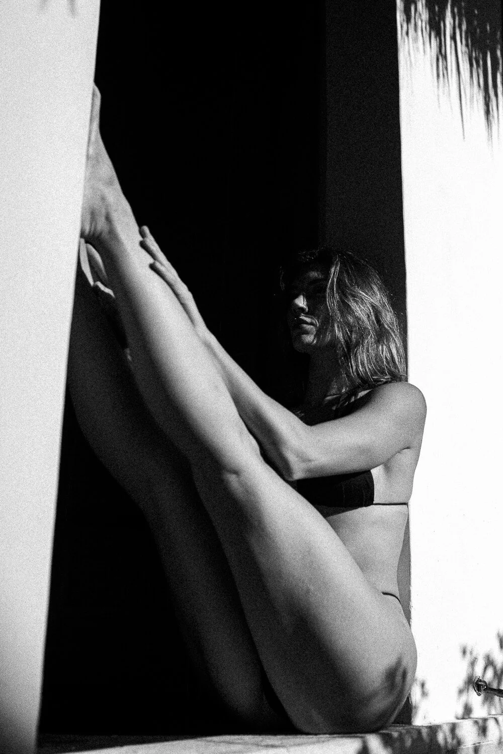 Black and white photo of a woman with curly hair wearing black lingerie, sitting on a surface with her legs extended in front of her, touching a wall or canvas with her hand, with shadows creating a dramatic effect.