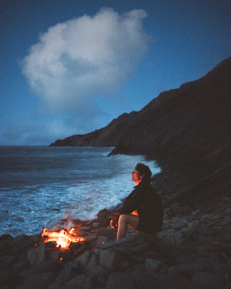 Une personne assise sur des rochers au bord de la mer, près d’un feu de camp, avec une falaise en arrière-plan et un ciel nocturne avec des nuages.
