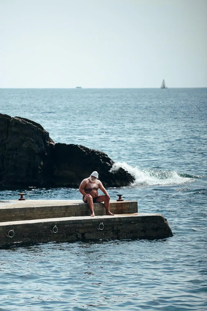 An elderly man in a white hat sits on the edge of a concrete dock by the water, with a rocky shoreline and the ocean behind him. Two sailboats are visible in the distance on the water.