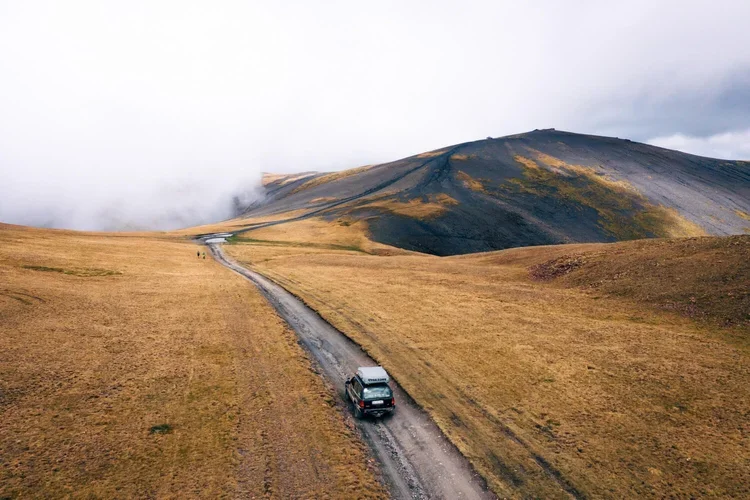 Un véhicule rouge circule sur un chemin de terre à travers un paysage de plaines dorées avec une montagne sombre en arrière-plan, sous un ciel nuageux.