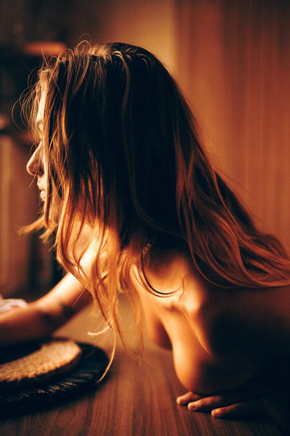 A woman with long, wavy hair in a warm, dimly lit room is leaning forward with her elbow on a table, with her head bowed and hair falling over her face.