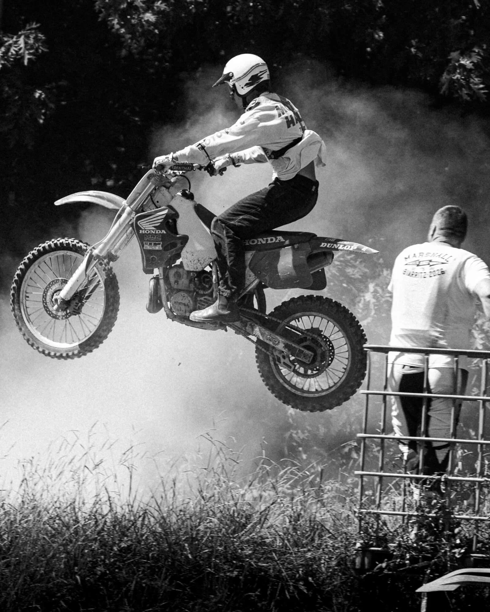 A person riding a dirt bike on a trail with dust in the air, wearing a helmet and protective gear, with another person standing nearby in a field of tall grass.