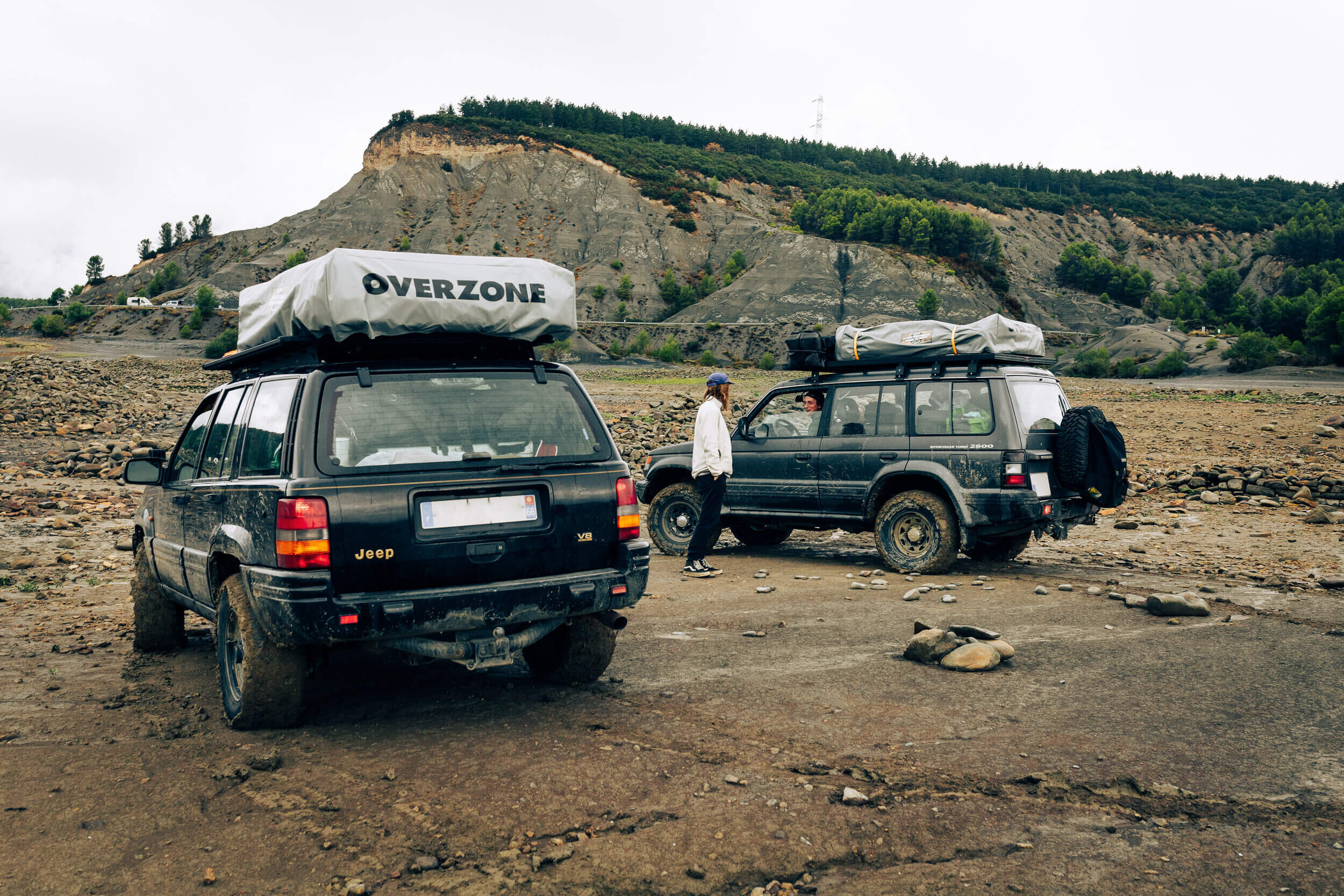 Deux SUV dans un paysage montagneux, avec des coffres de toit chargés. Une personne se tient près de l'un des véhicules.