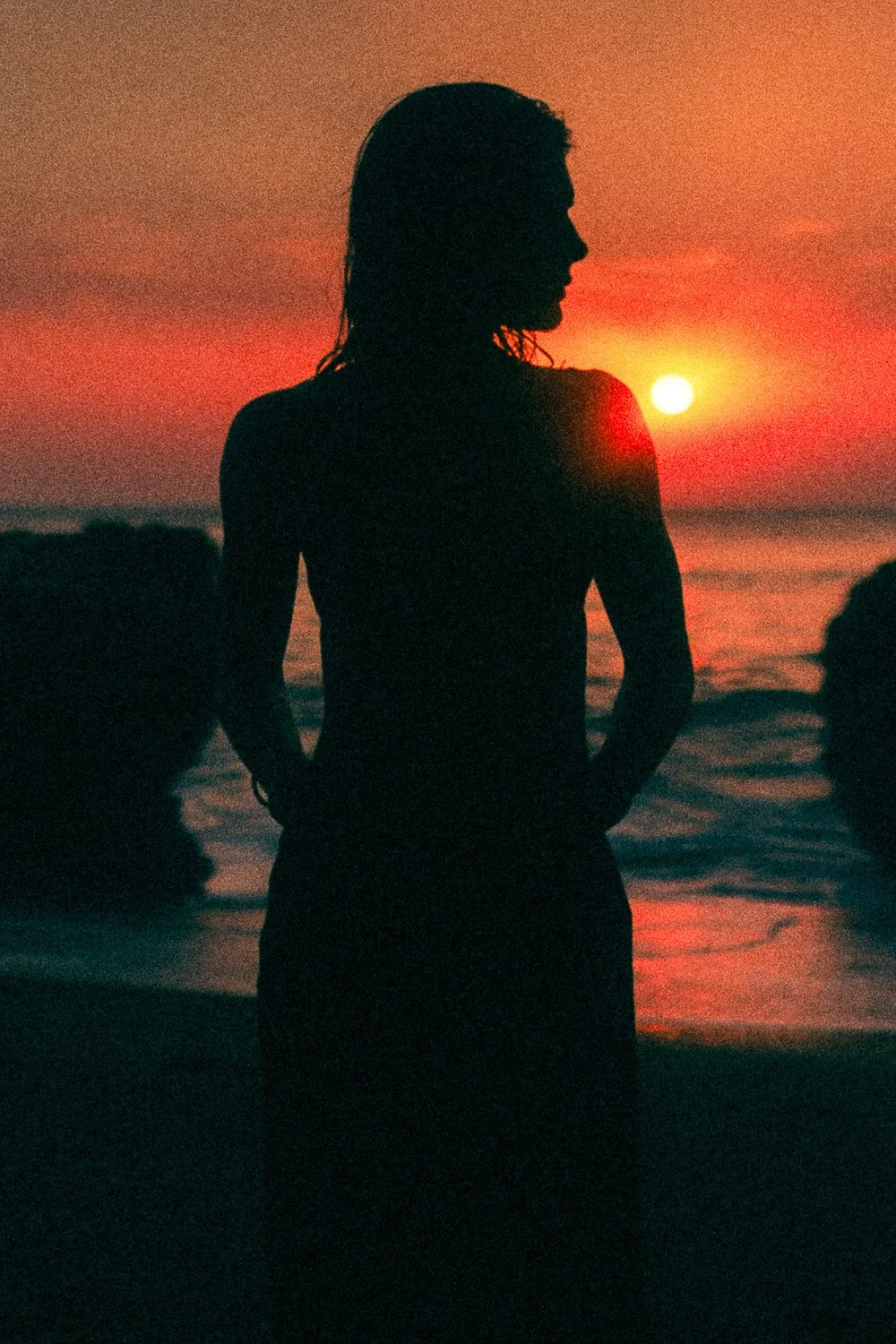 Silhouette of a woman standing on the beach during sunset, with the sun near the horizon and waves in the background.