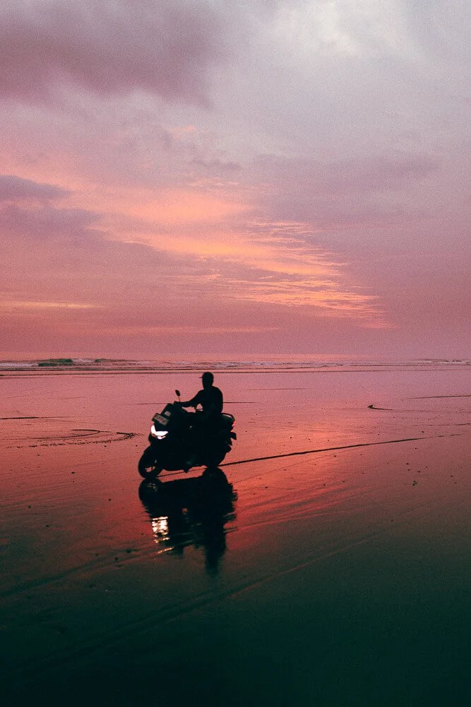 Person riding a scooter on a wet beach during sunset with pink and purple sky.