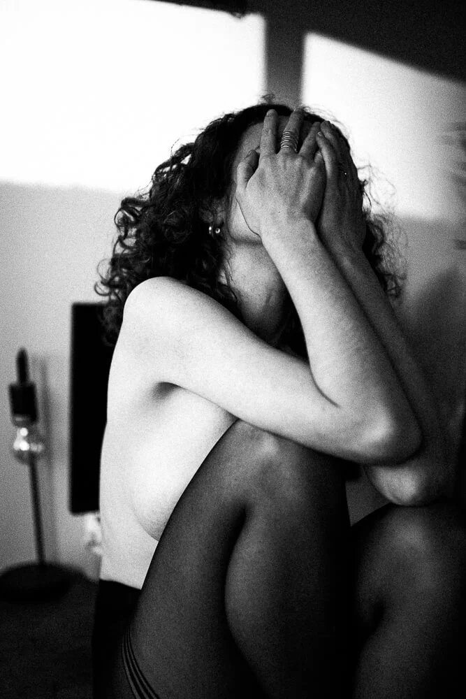 A woman with curly hair covering her face with her hands, sitting with her knees up, in a black-and-white photo.
