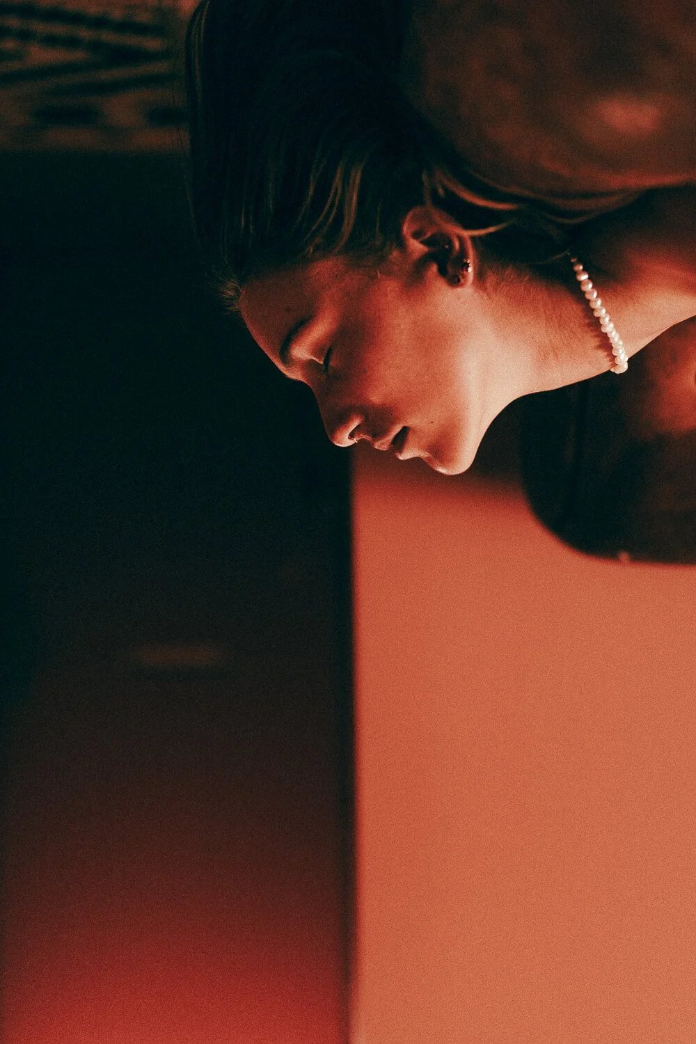 A young woman with long dark hair, wearing pearl earrings and a pearl necklace, is looking downward with her eyes closed, illuminated by warm, soft lighting.